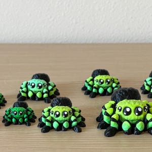 May include: A collection of ten small, three-dimensional spider figurines in various sizes. Most spiders feature a bright lime green and black striped pattern with large, expressive eyes. One spider is brown with tan markings, and one is purple.