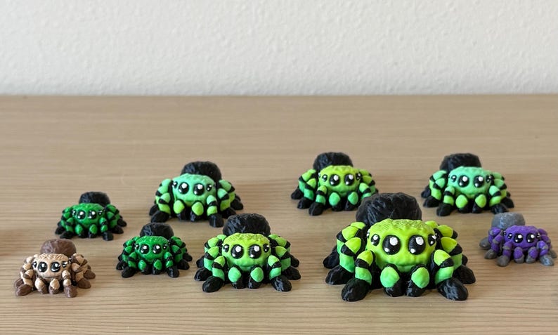 May include: A collection of ten small, three-dimensional spider figurines in various sizes. Most spiders feature a bright lime green and black striped pattern with large, expressive eyes. One spider is brown with tan markings, and one is purple.