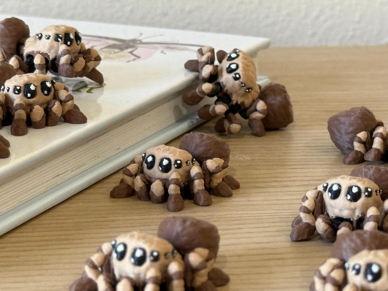May include: A collection of small, sculpted spider figurines featuring a tan and dark brown color palette. Each spider has large, expressive black eyes with white highlights, a rounded abdomen, and multiple segmented legs in a crouching pose.