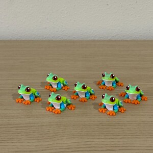 May include: Seven small, colorful frog figurines. Each frog has a green body, blue sides, orange feet, and red eyes. They are arranged on a light brown wooden surface.