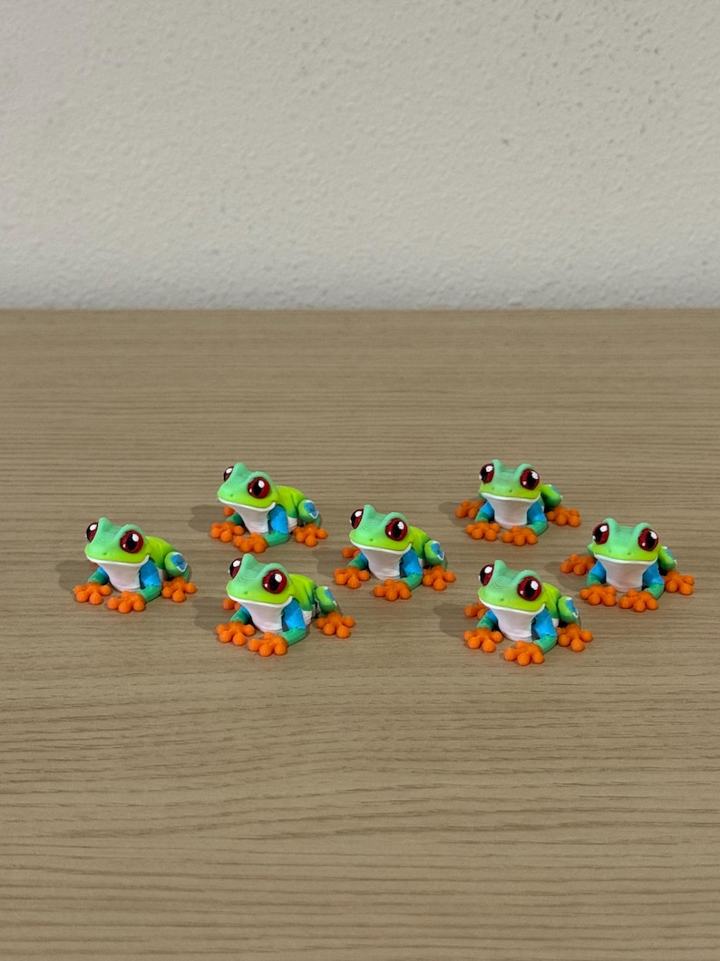 May include: Seven small, colorful frog figurines. Each frog has a green body, blue sides, orange feet, and red eyes. They are arranged on a light brown wooden surface.