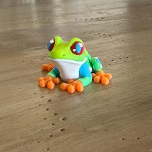May include: A colorful 3D-printed frog figurine. The frog features a vibrant green head, blue body, and orange feet. Its eyes are red with blue and white accents. The frog is sitting on a wooden surface.