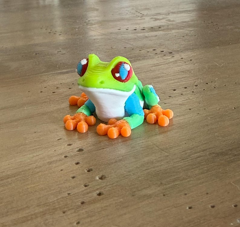 May include: A colorful 3D-printed frog figurine. The frog features a vibrant green head, blue body, and orange feet. Its eyes are red with blue and white accents. The frog is sitting on a wooden surface.