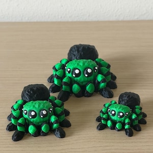 May include: Three spider figurines in varying sizes, each featuring a bright green body with black accents and large, expressive white-dotted eyes. The spiders have textured black abdomens and multiple legs, arranged in a group on a flat surface.