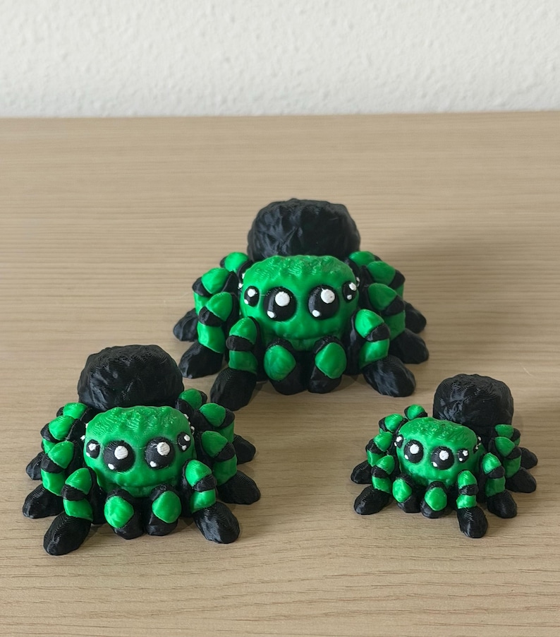 May include: Three spider figurines in varying sizes, each featuring a bright green body with black accents and large, expressive white-dotted eyes. The spiders have textured black abdomens and multiple legs, arranged in a group on a flat surface.