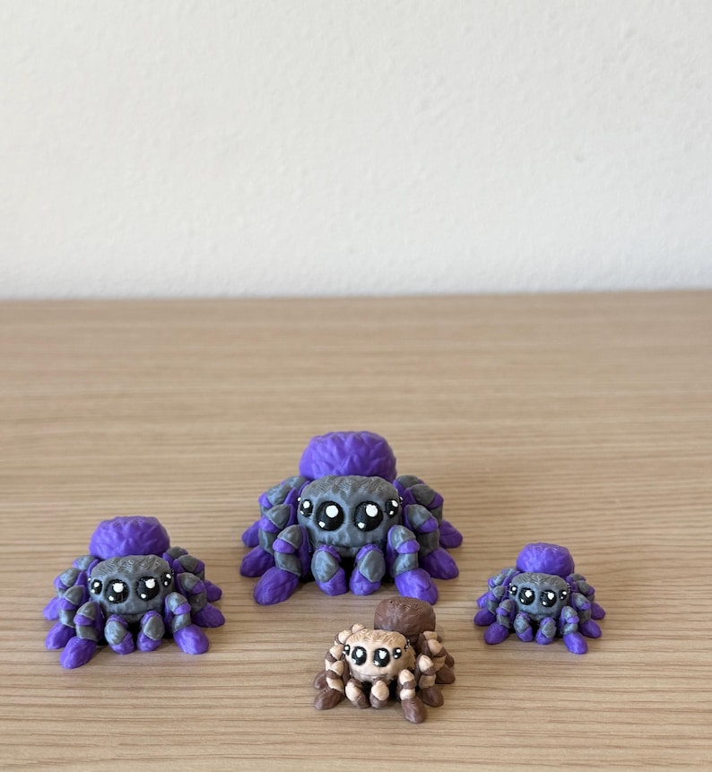 May include: A collection of four small spider figurines. Three spiders are purple and gray with large black eyes and white highlights. One smaller spider is brown and tan with similar large black eyes and white highlights, positioned in the front.