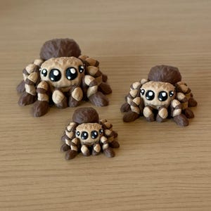 May include: Three brown and tan spider figurines of varying sizes, each featuring a rounded body, multiple textured legs, and large, expressive black eyes with white highlights. The spiders are arranged in a group, showcasing their detailed, sculpted forms and consistent color palette across all three pieces.
