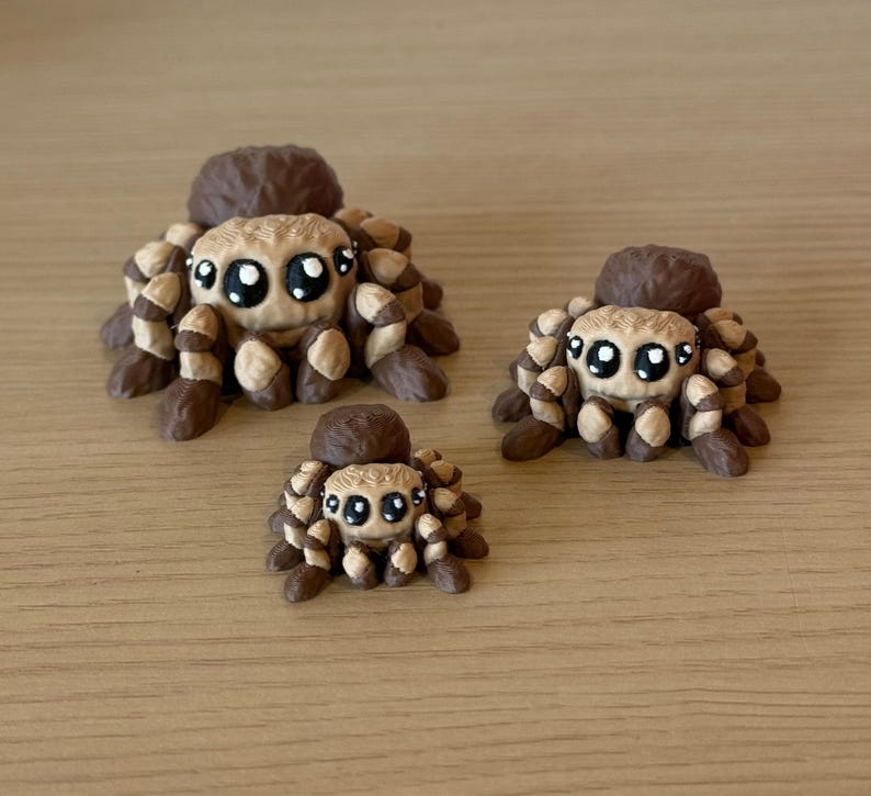 May include: Three brown and tan spider figurines of varying sizes, each featuring a rounded body, multiple textured legs, and large, expressive black eyes with white highlights. The spiders are arranged in a group, showcasing their detailed, sculpted forms and consistent color palette across all three pieces.