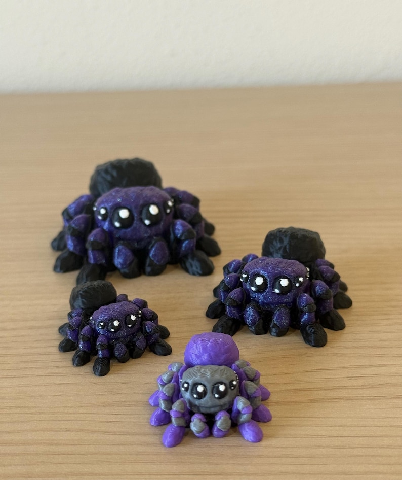 May include: A collection of four small spider figurines with large eyes. Three spiders are purple with black legs and abdomens, while the smallest spider is light purple and gray with a purple abdomen. Each spider has eight legs and a rounded body.