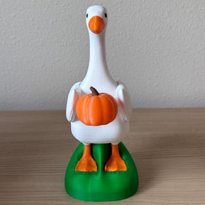 Puede incluir: Una figura de ganso blanco con pico y patas naranjas, de pie sobre una base verde. El ganso sostiene una peque&ntilde;a calabaza naranja redonda con un tallo marr&oacute;n entre sus alas. La figura tiene un acabado mate y se mantiene en posici&oacute;n vertical.