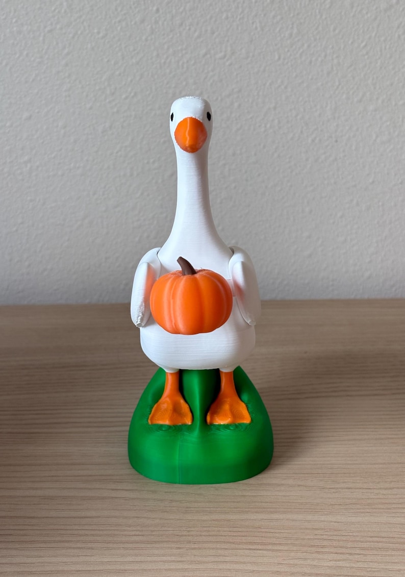 Puede incluir: Una figura de ganso blanco con pico y patas naranjas, de pie sobre una base verde. El ganso sostiene una peque&ntilde;a calabaza naranja redonda con un tallo marr&oacute;n entre sus alas. La figura tiene un acabado mate y se mantiene en posici&oacute;n vertical.