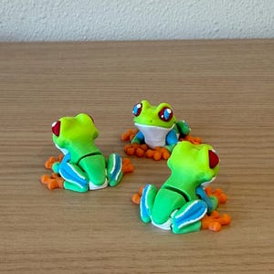 May include: Three handmade frog figurines. Each frog is lime green with red eyes, blue accents, and orange feet. The frogs are sitting on a wooden surface.