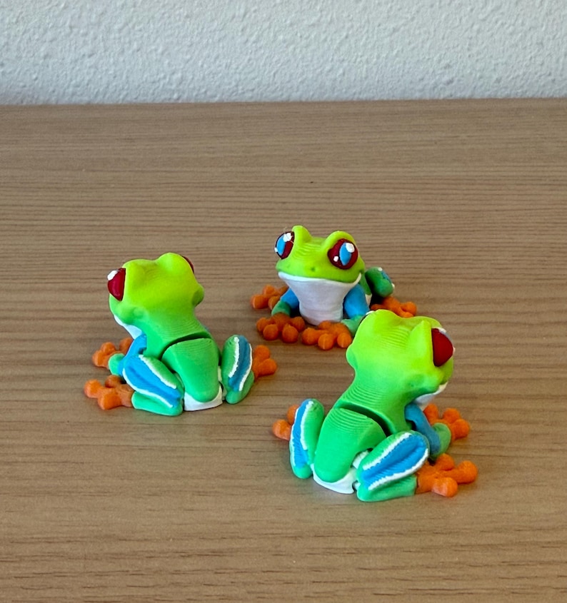 May include: Three handmade frog figurines. Each frog is lime green with red eyes, blue accents, and orange feet. The frogs are sitting on a wooden surface.