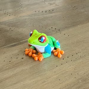 May include: A colorful frog figurine with a green head, back, and blue accents. The frog has orange feet and large eyes with red, white, and blue details. The figurine is sitting on a wooden surface.