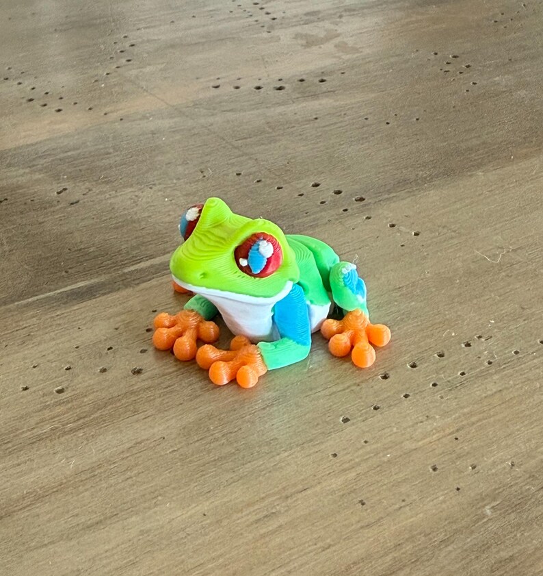 May include: A colorful frog figurine with a green head, back, and blue accents. The frog has orange feet and large eyes with red, white, and blue details. The figurine is sitting on a wooden surface.