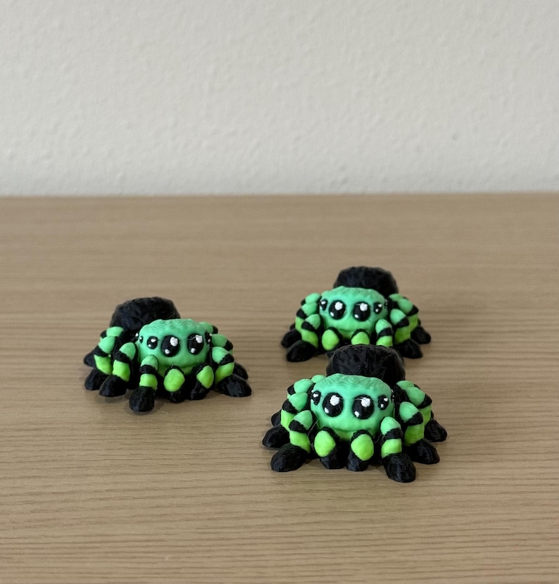 May include: Three small spider figurines featuring a bright light green body with black striped legs and large, dark, glossy eyes with white highlights. Each spider has a rounded, textured appearance and is positioned in a cluster.