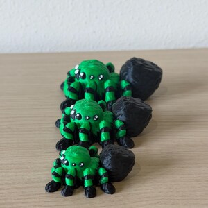 May include: Three 3D printed spider figurines arranged in a line from largest to smallest. Each spider features a bright green body with black striped legs and a large, textured black abdomen. They have multiple white eyes on their heads.