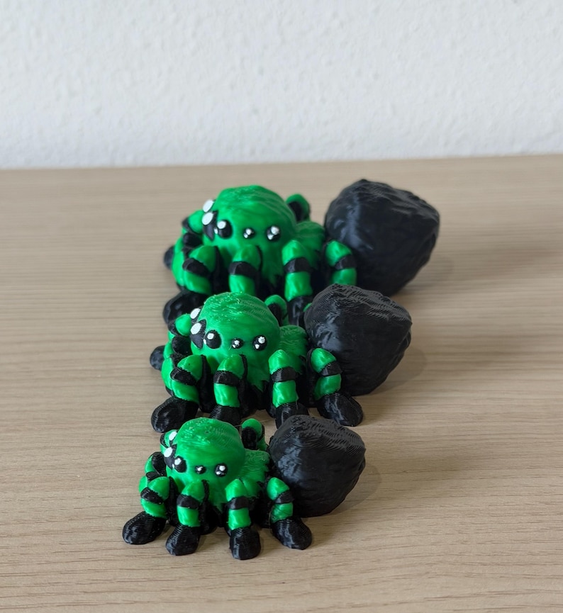 May include: Three 3D printed spider figurines arranged in a line from largest to smallest. Each spider features a bright green body with black striped legs and a large, textured black abdomen. They have multiple white eyes on their heads.
