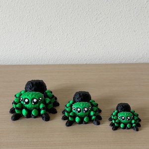 May include: Three spider figurines in descending sizes, each featuring a bright green body with black accents and large, round white eyes. The spiders have a textured, multi-faceted surface and are arranged in a row from largest to smallest.