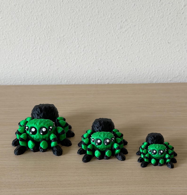 May include: Three spider figurines in descending sizes, each featuring a bright green body with black accents and large, round white eyes. The spiders have a textured, multi-faceted surface and are arranged in a row from largest to smallest.
