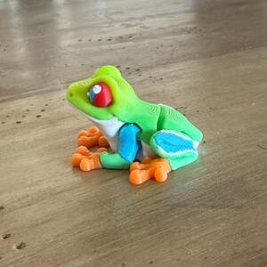 May include: A 3D-printed frog figurine in bright green, blue, orange, and white. The frog has large red and white eyes and orange feet. The figurine is sitting on a wooden surface.