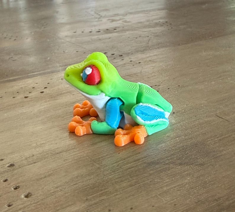 May include: A 3D-printed frog figurine in bright green, blue, orange, and white. The frog has large red and white eyes and orange feet. The figurine is sitting on a wooden surface.