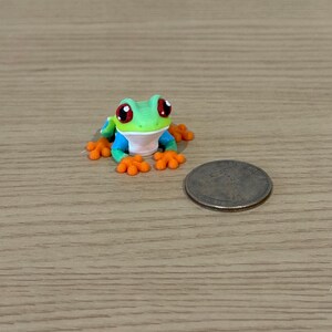 May include: A miniature, colorful frog figurine with large red eyes, green head, blue arms, and orange feet. A coin is next to the frog. The frog is on a wooden surface.