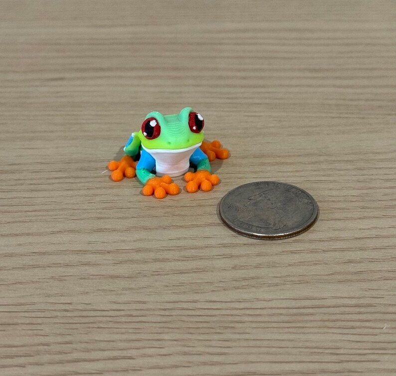 May include: A miniature, colorful frog figurine with large red eyes, green head, blue arms, and orange feet. A coin is next to the frog. The frog is on a wooden surface.