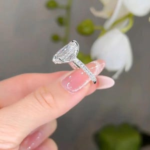 May include: A silver ring with a large, pear-shaped diamond center stone. The band is encrusted with smaller diamonds. The ring is held up against a blurred background of white flowers and green foliage.