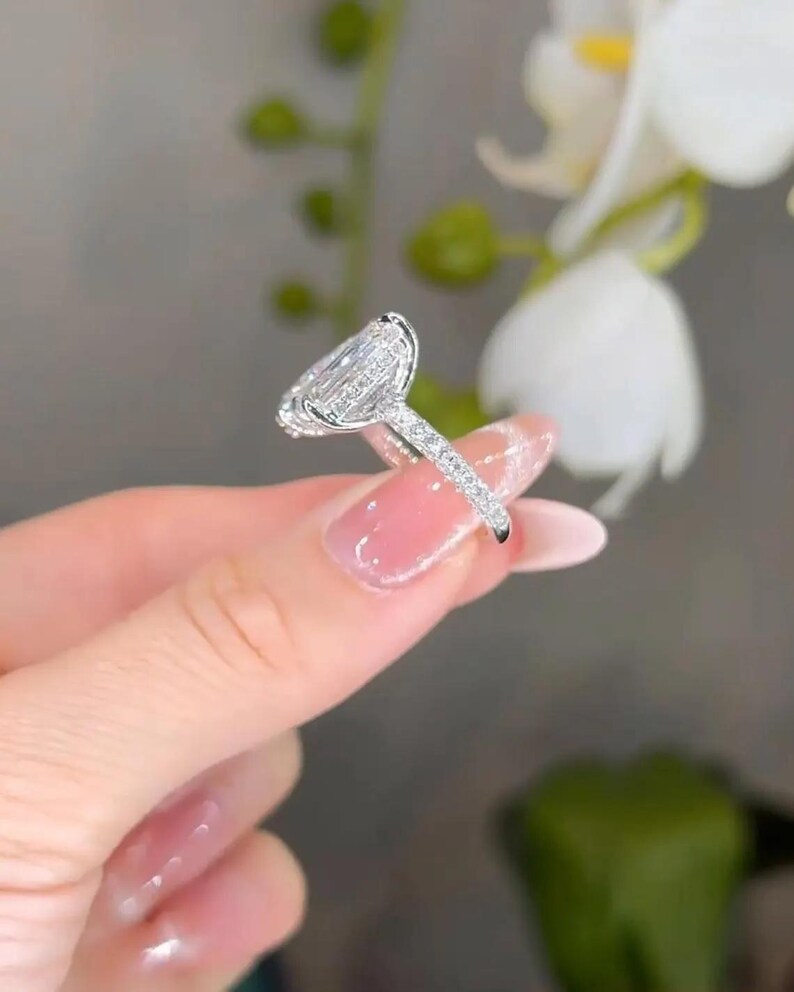 May include: A silver ring with a large, pear-shaped diamond center stone. The band is encrusted with smaller diamonds. The ring is held up against a blurred background of white flowers and green foliage.
