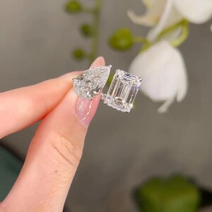 May include: Two diamond rings, one with a pear-shaped stone and the other with an emerald-cut stone. Both rings are set in a silver band. The rings are held up against a blurred background of white flowers.