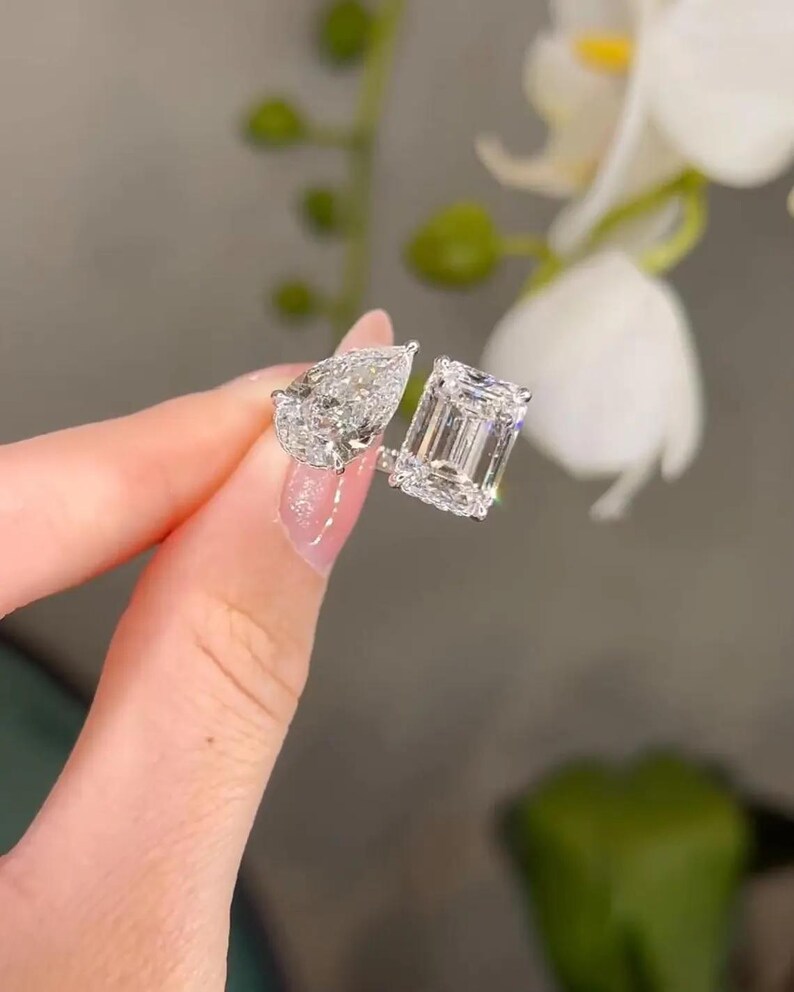 May include: Two diamond rings, one with a pear-shaped stone and the other with an emerald-cut stone. Both rings are set in a silver band. The rings are held up against a blurred background of white flowers.