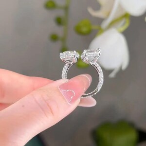 May include: A silver ring with two large, clear, square-cut stones. The ring band is encrusted with small, sparkling stones. The ring is held up against a soft-focus background of white flowers and green foliage.