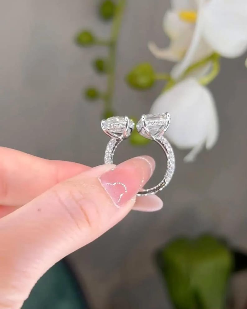 May include: A silver ring with two large, clear, square-cut stones. The ring band is encrusted with small, sparkling stones. The ring is held up against a soft-focus background of white flowers and green foliage.