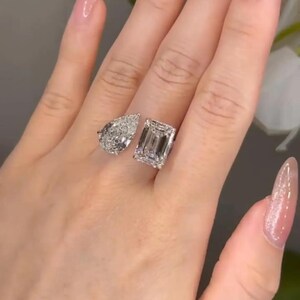 May include: Two diamond rings, one pear-shaped and one emerald-cut, are displayed. The rings are set on a hand with long, polished nails. The diamonds sparkle, reflecting light.