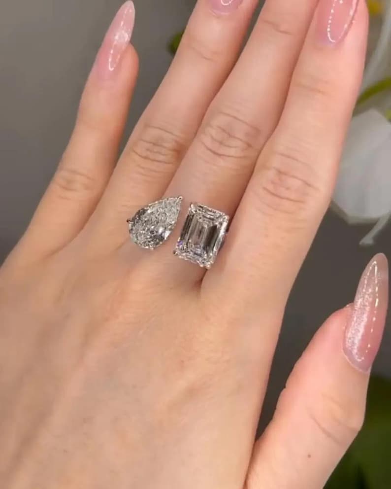 May include: Two diamond rings, one pear-shaped and one emerald-cut, are displayed. The rings are set on a hand with long, polished nails. The diamonds sparkle, reflecting light.