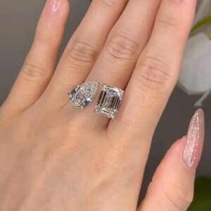 May include: Two diamond rings, one pear-shaped and one emerald-cut, are displayed on a hand. The rings sparkle with brilliant facets, reflecting light. The hand has long, manicured nails with a light pink polish.