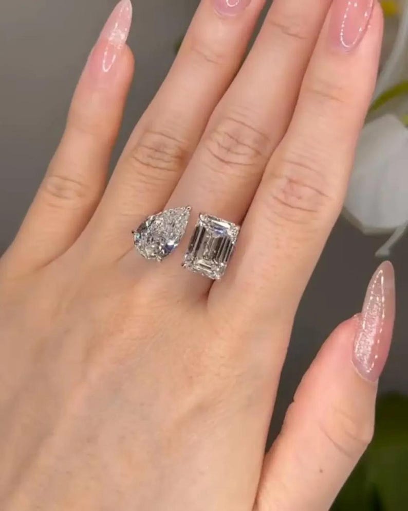 May include: Two diamond rings, one pear-shaped and one emerald-cut, are displayed on a hand. The rings sparkle with brilliant facets, reflecting light. The hand has long, manicured nails with a light pink polish.