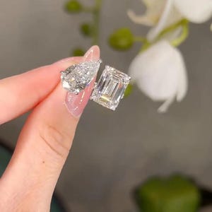 May include: Two diamond earrings, one pear-shaped and one emerald-cut, are held up against a blurred floral background. The earrings sparkle, reflecting light. The setting appears to be silver or white gold.