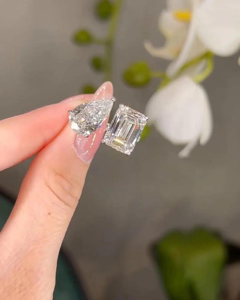 May include: Two diamond earrings, one pear-shaped and one emerald-cut, are held up against a blurred floral background. The earrings sparkle, reflecting light. The setting appears to be silver or white gold.