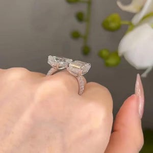 May include: Two silver rings with large, rectangular, clear gemstones. The rings are held up, showcasing the sparkling stones and the delicate, gem-studded bands. The background is blurred, highlighting the jewelry.