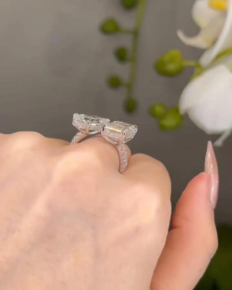 May include: Two silver rings with large, rectangular, clear gemstones. The rings are held up, showcasing the sparkling stones and the delicate, gem-studded bands. The background is blurred, highlighting the jewelry.