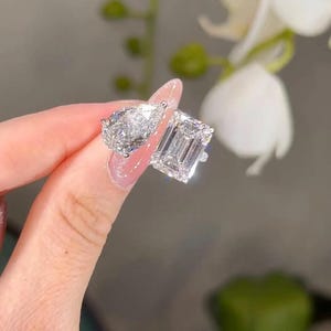 May include: Two diamond rings, one pear-shaped and one emerald-cut, sparkle brightly. The rings are held up, showcasing their facets and brilliance. The setting appears to be a light-colored metal, enhancing the diamonds' clarity.