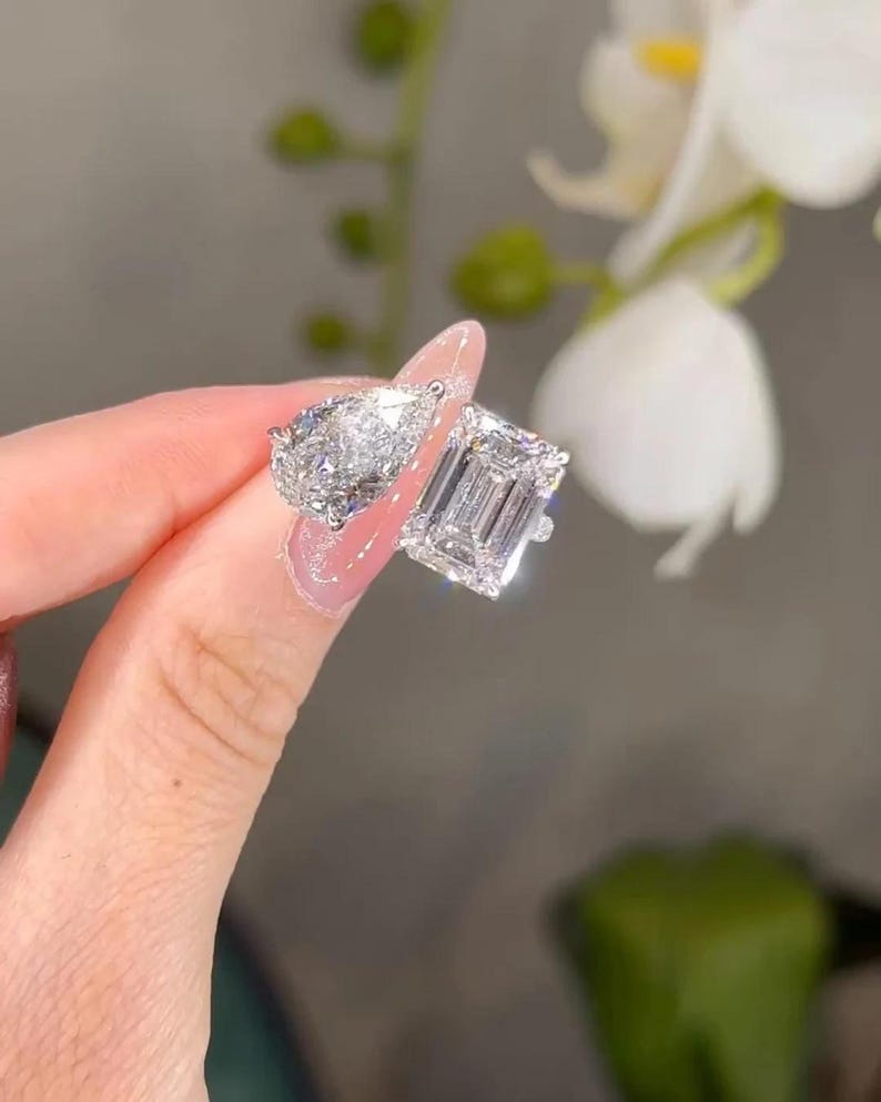 May include: Two diamond rings, one pear-shaped and one emerald-cut, sparkle brightly. The rings are held up, showcasing their facets and brilliance. The setting appears to be a light-colored metal, enhancing the diamonds' clarity.