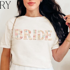 Puede incluir: Camiseta color marfil con la palabra "BRIDE" en un estampado floral. La camiseta se combina con una falda plisada blanca. La palabra "IVORY" está impresa en la esquina superior izquierda. Un bolso negro cuelga de una cadena. Tema nupcial.
