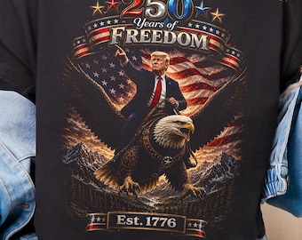 May include: Black sweatshirt featuring a graphic of a man riding an eagle in front of an American flag, with the text "250 Years of Freedom" and "Est. 1776". The sweatshirt is worn by a person.