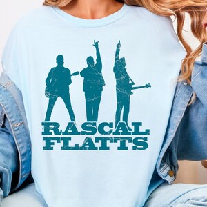May include: Light blue t-shirt featuring a teal graphic of three musicians and the text "RASCAL FLATTS". The shirt is made of a soft material and is suitable for casual wear. The shirt is a crew neck style.