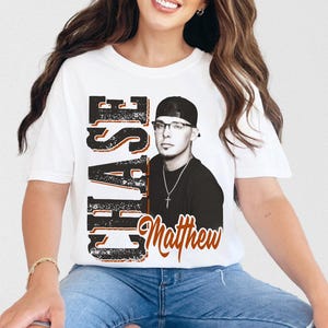 May include: White t-shirt featuring a graphic of a man wearing glasses and a baseball cap. The shirt has the name "CHASE" in large, distressed black letters with orange accents, and the name "Matthew" in orange script.