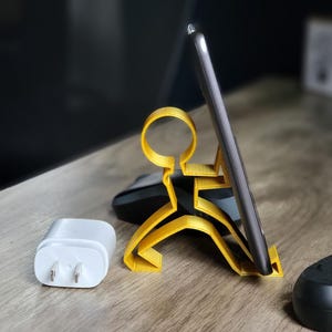 May include: A gold-colored 3D printed phone stand shaped like a stick figure holding a phone. The phone stand is on a wooden surface with a white phone charger and a black object in the background.