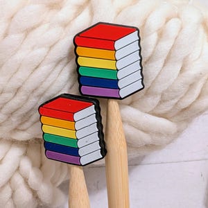 May include: Two book-shaped charms with rainbow-colored pages and red covers. The charms are attached to wooden sticks, set against a backdrop of thick, white yarn. The books are a vibrant display of color.